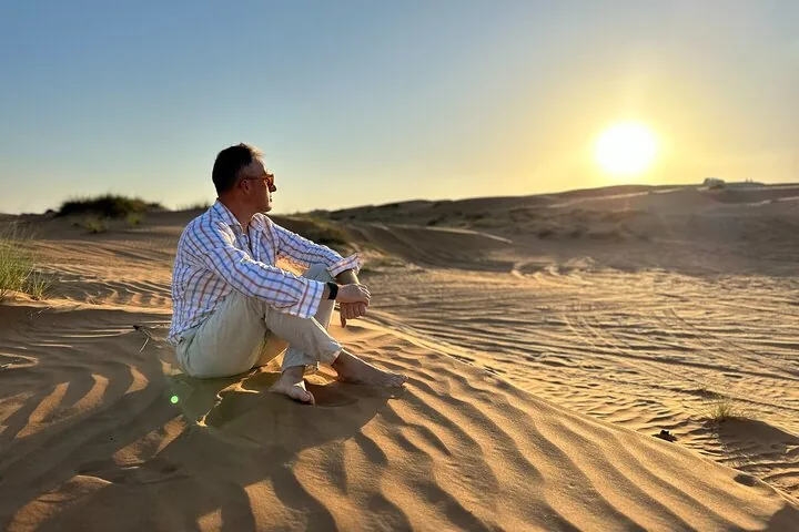 A man sitting on a sand dune looking at the setting sun.